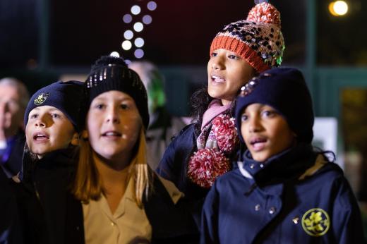 A group of children in coats and hats singing and looking up at the lights