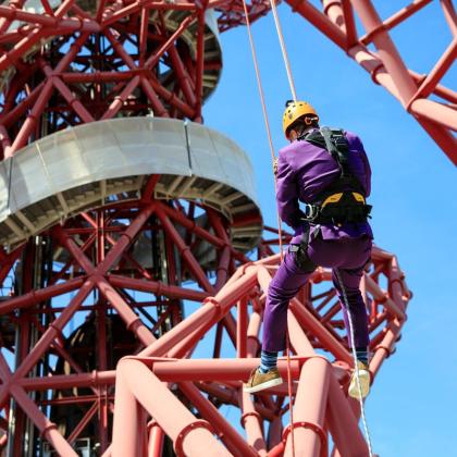 A man in a purple suit and orange helmet abseils from the Accelormittal Sculpture in Olympic Park, London
