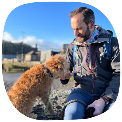 A man sitting on a beach, wearing a winter jacket and stroking a small fluffy ginger dog
