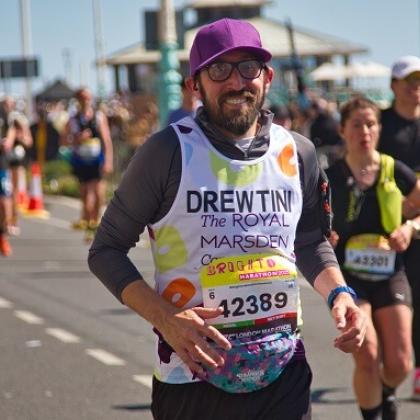 Royal Marsden runner Andrew smiling at camera with a purple baseball hat and glasses on. There are other runners blurred in the background.