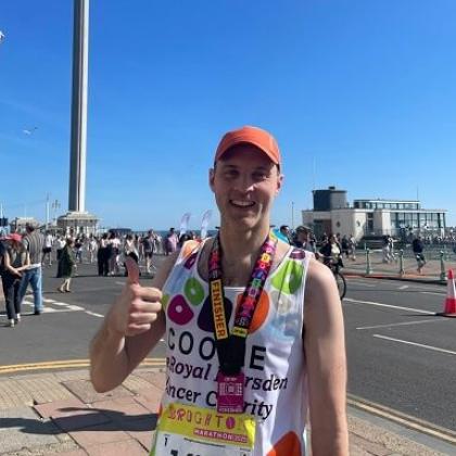 Royal Marsden runner smiling with medal, standing in front of the Brighton i360. He has a proud smile on his face, there's blue skies in the background and he's wearing an orange hat.