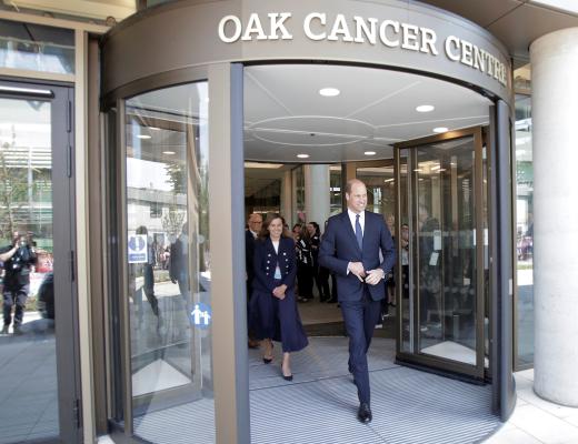 His Royal Highness Prince William dressed smartly in a suit walking out of large glass sliding doors of the Oak Cancer Centre 