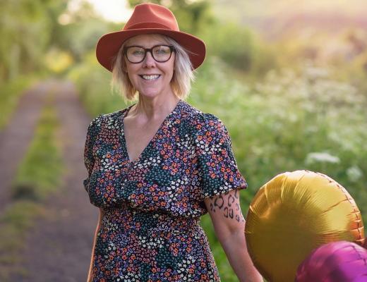 Royal Marsden patient Julie in a floral dress and hat, standing on a country path in the summer, smiling and holding balloons