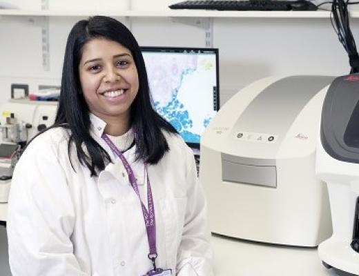 A Biomedical Scientist in a white lab coat smiling and standing with stainers and a piece of white equipment in a hospital medical room