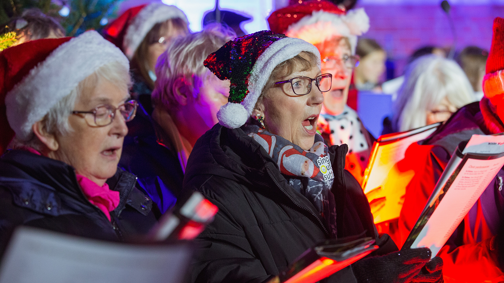 A colourful photo of group of ladies singing carols, wearing santa hats at a Celebrate a life carol service. 