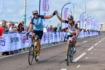 A couple cycling over the London to Brighton Cycle Ride finish line. The are holding hands in the air in celebration and wearing Royal Marsden Cancer Charity cycling vests