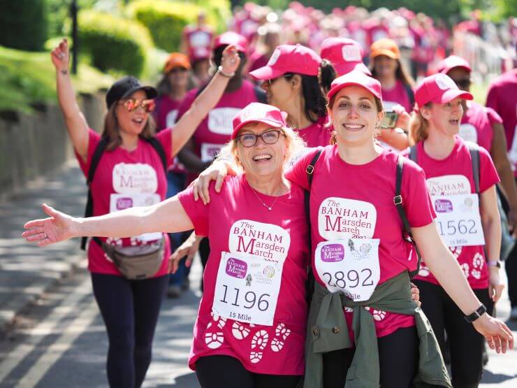 Two ladies wearing pink t-shirts, caps, and fundraising event numbers smile at the finish line of the Banham Marsden March