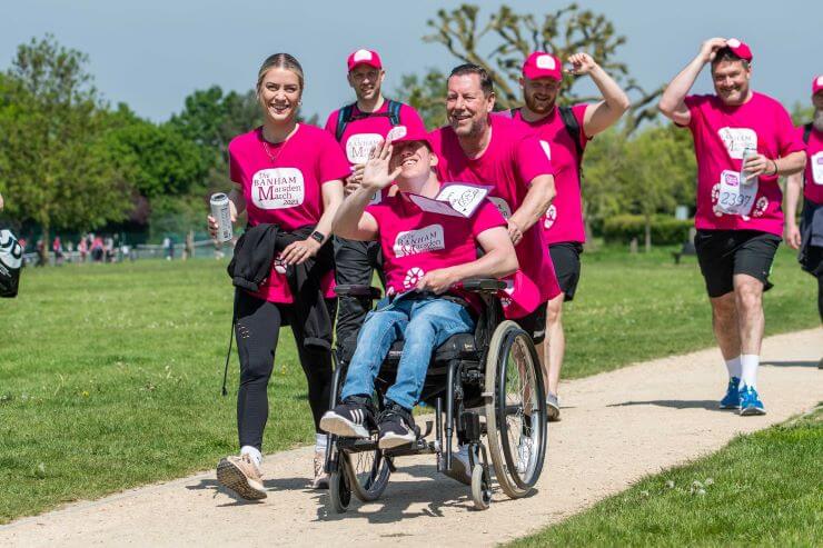 A group of six fundraisers wearing bright pink Banham Marsden March t-shirts. They are smiling and walking along a path together. One of the fundraisers at the front is smiling and waving at the camera - he is in a wheelchair. 