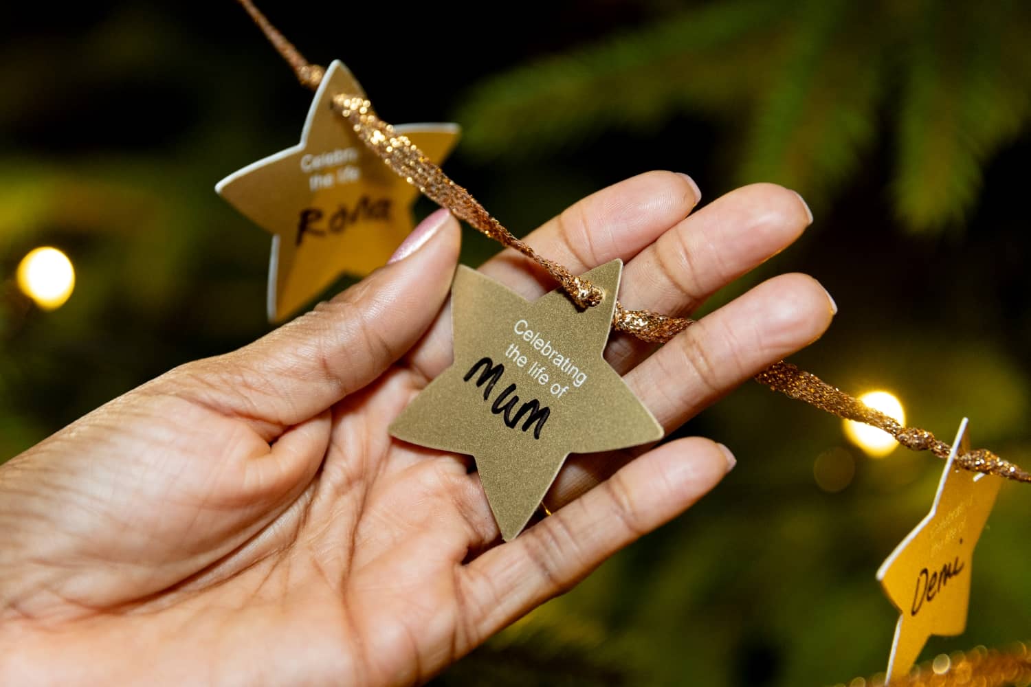 A close up photo of a hand holding a golden star decoration, hung on a christmas tree. The star says 'Celebrating the life of Mum' with the word 'mum' handwritten on the star.