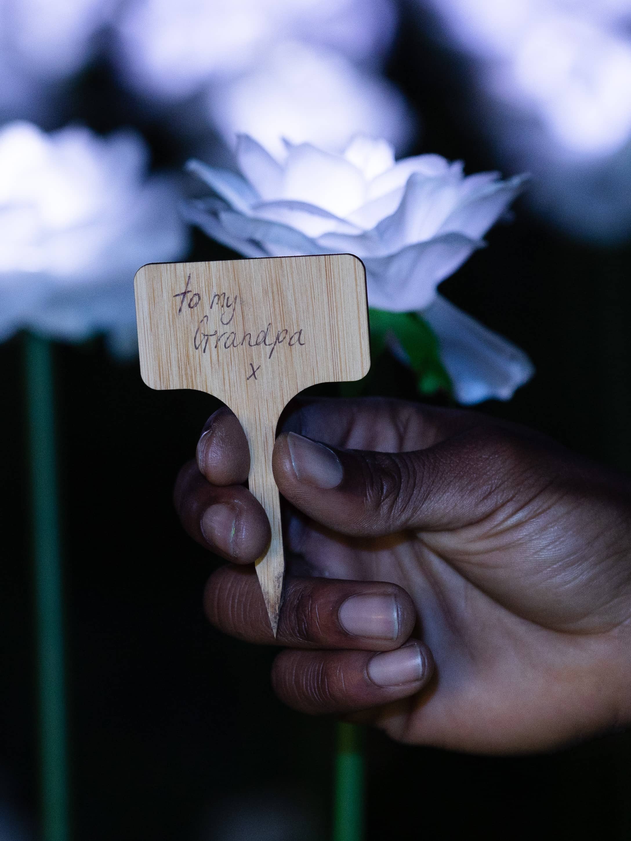 A hand holding a small wooden dedication sign. The handwritten dedication reads 'To my Grandpa x'. The illuminated roses of the Ever After Garden are visible in the background