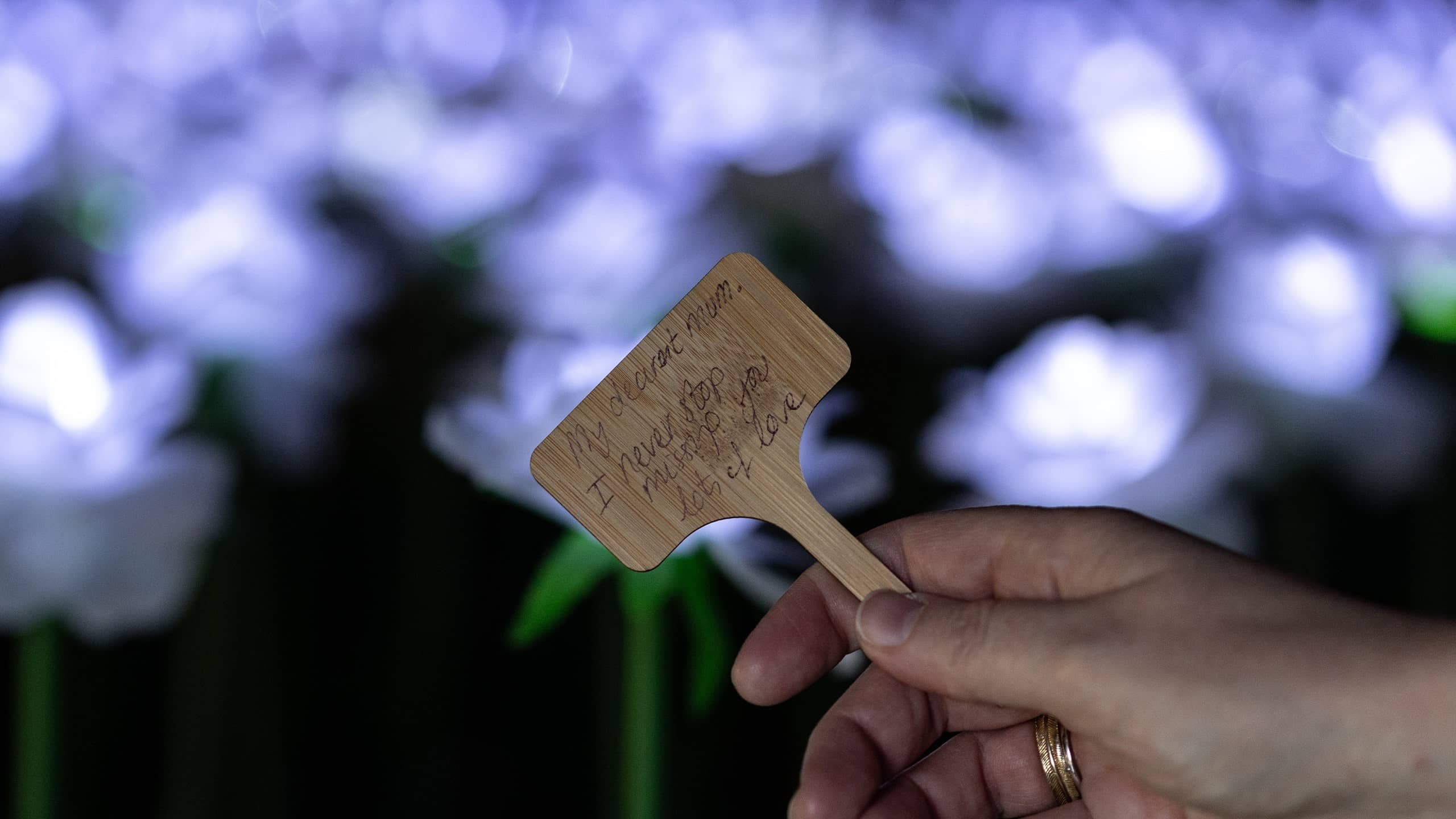 A photo of a hand holding a small wooden sign. The sign has a dedication written on it which reads - 'My dearest Mum, I never stop missing you. Lots of love' handwritten on it.