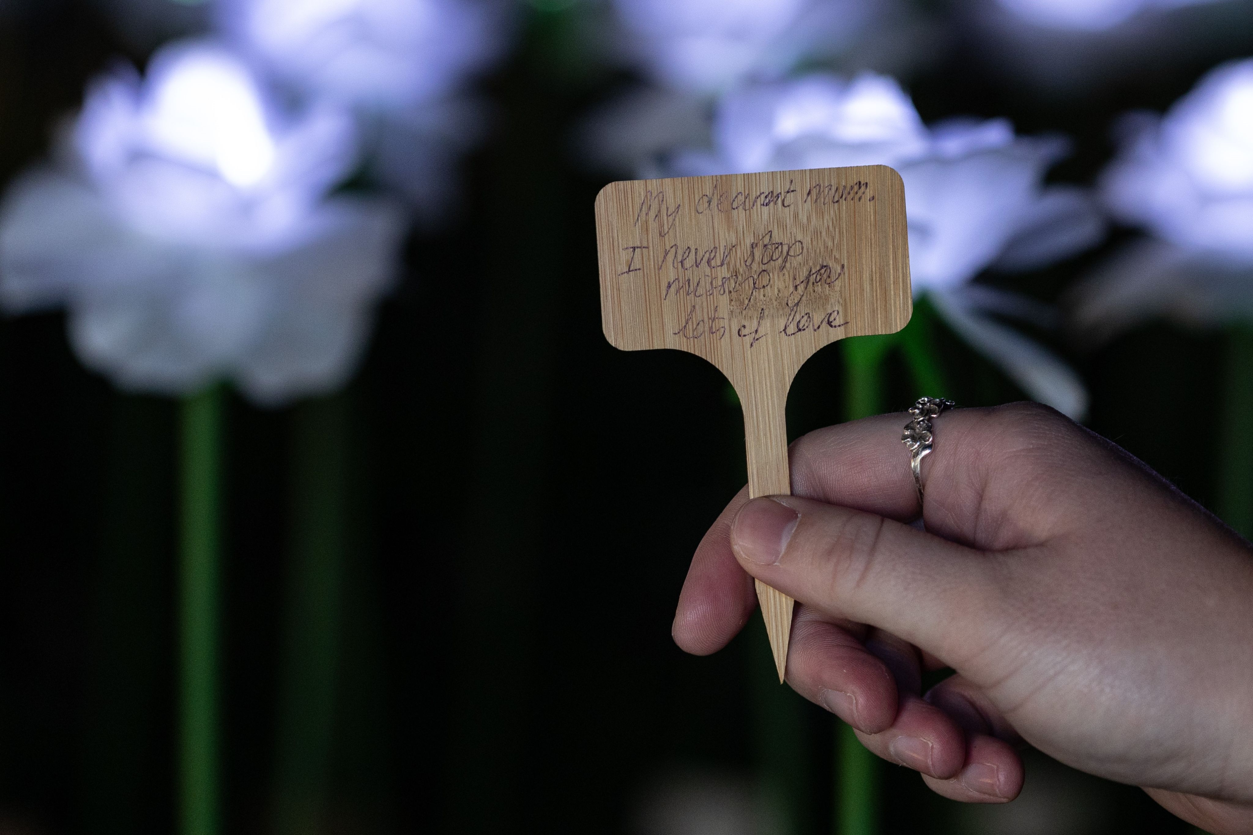 A photo of a hand holding small wooden sign. Th sign has a dedication written on it which reads - 'My dearest Mum, I never stop missing you. Lots of love' handwritten on it.
