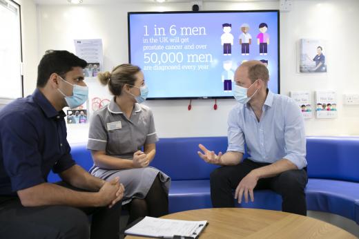 A dark-haired man sits next to a woman in a nurse's uniform, they are talking to HRH The Duke of Cambridge who is sat opposite them. There is a digital screen behind them, all are wearing surgical masks