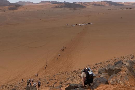 Landscape photo of runners in the desert. They are approaching a checkpoint which is far in the distance. Mountains are in the background