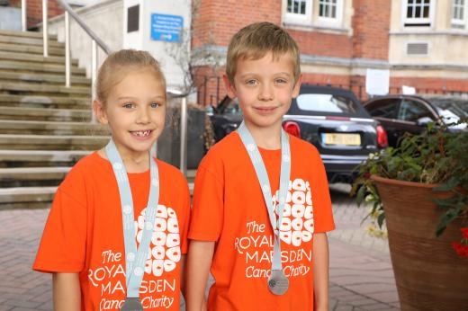 Reece and Emilie outside The Royal Marsden hospital in Chelsea. They are smiling, wearing orange charity branded t-shirts and have medals 
