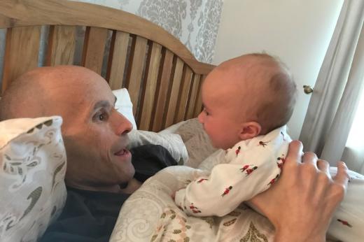 Simon laying in bed holding up baby Dexter. They are looking at each other and smiling.