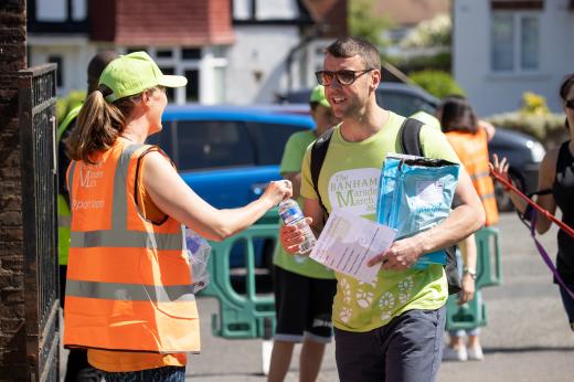 A young woman in a Banham Marsden March orange volunteer vest and cap hands out a water to someone taking part in the March. 