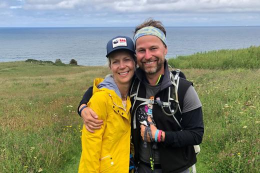 Young man and woman arm in arm on the South downs. They are wearing hiking gear and smiling at the camera. The man has a Royal Marsden Cancer Charity shirt on and the woman wears a bright yellow jacket. 