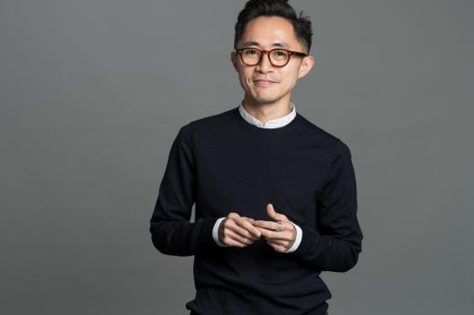 Ken standing infront of a grey background. He is wearing a dark sweatshirt and brown rimmed classes.