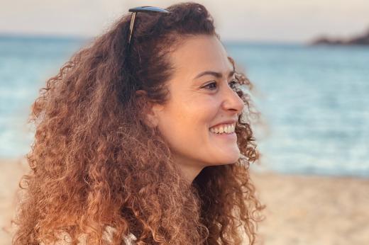 A photo of Klodjana. She has a big smile and brown curly hair. She is on a beach.