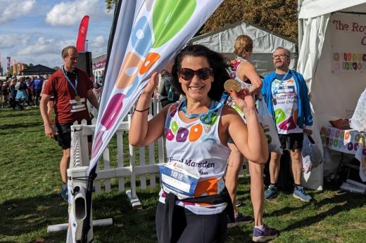 Klodjana after completing the Royal Parks half Marathon. She is standing by a Royal Marsden Cancer Charity banner and holding up her marathon medal. She has a big smile.