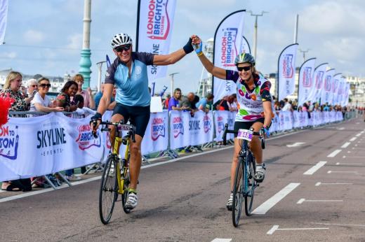 Chris and Carolyn Cycling over the London to Brighton Cycle Ride finish line. They are smiling and holing each others hands in the air in celebration. They are wearing Royal Marsden Cancer Charity cycling vests