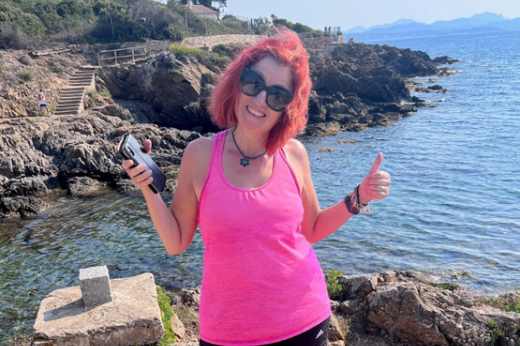 A photo of Pip. She is outside standing in from of a rocky shoreline. She has a big smile and is wearing a bright pink running shirt.