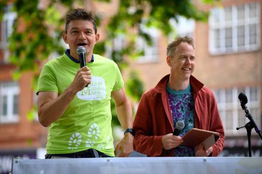 A speaker in a Banham Marsden March T-shirt speaking at the start line. he is smiling and holding a microphone. 