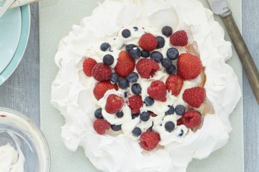 A white meringue nest with cream, raspberries, blueberries and strawberries in the middle, on a serving board 