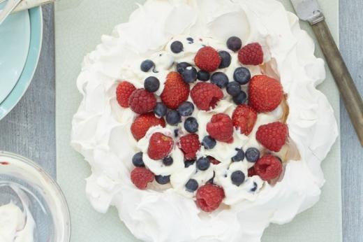 A white meringue nest with cream, raspberries, blueberries and strawberries in the middle, on a serving board