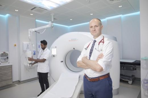 Dr Lee standing in front of a CT Scanner and a radiographer behind him using the machine