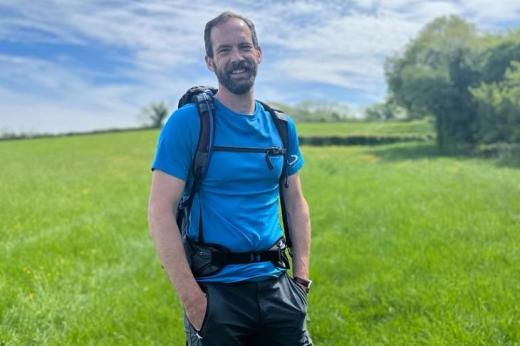 Jon standing in a rolling green field on a sunny day, wearing a backpack and walking gear.