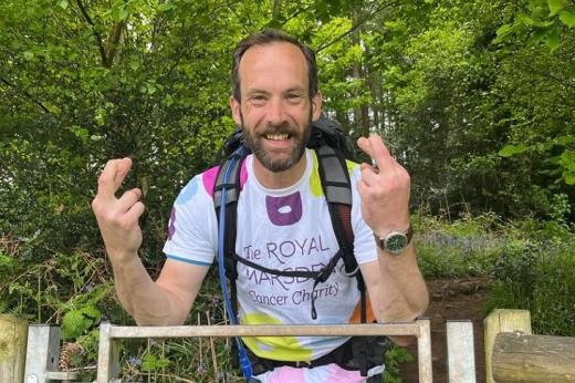 Jon leaning on a fence in a wooded area, wearing a backpack, walking gear and a Royal Marsden Cancer Charity t-shirt