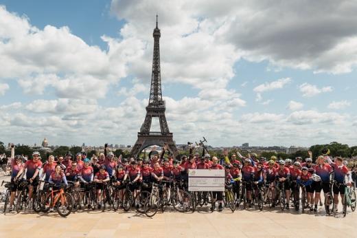 A large group of cyclists dismounted from their bikes, standing and waving in front of the Eiffel tower