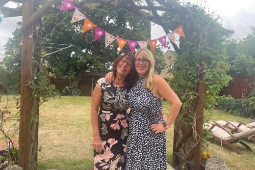 Two women in summer dresses standing under a garden trellis with Royal Marsden Cancer Charity bunting tied to it