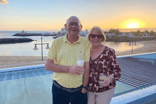 Two people holding drinks and standing on a rooftop overlooking a sandy beach at sunset