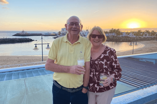 Two people holding drinks and standing on a rooftop overlooking a sandy beach at sunset 