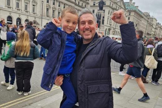 A man carrying his son on a busy street in London, throwing their fists in the air in happiness