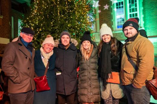 A group of six people in coats and hats standing in front of a large Christmas tree and projected star lights