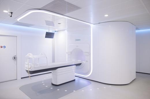 A white medical room with a large white machine in the middle, resembling an MRI with a flat bed and a tunnel