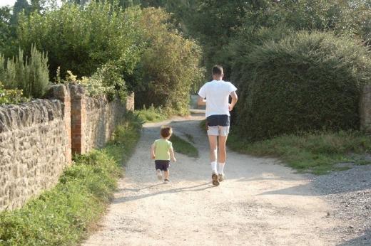 The back of a man in running gear jogging down a country road with a small toddler beside him