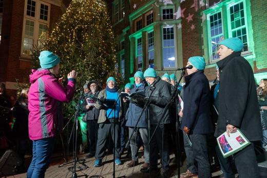 A choir group singing into microphones underneath a large Christmas tree decorated in stars and lights