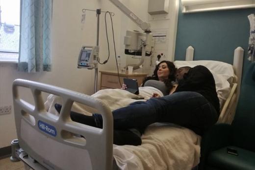 Two people lying together on a hospital bed in a hospital room, looking at a tablet device