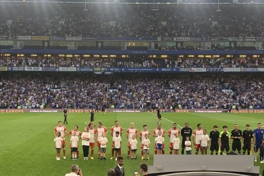 A football ground full of fans, with a line of players on the pitch and child mascots in front of them wearing RMCC t-shirts