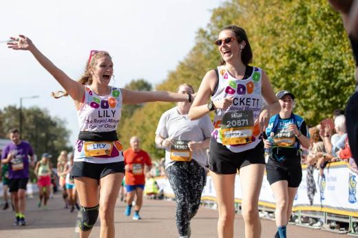 Two women smiling in Royal Marsden Cancer Charity gear, running down a road surrounded by other runners and people cheering