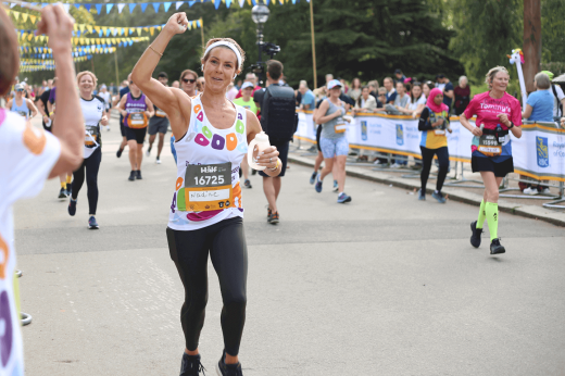 A runner in a Royal Marsden Cancer Charity t-shirt, first pumping and holding a water bottle as she jogs past supporters on the side of the road