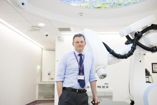 A smartly-dressed Royal Marsden doctor with a lanyard, standing next to a large white machine with a robotic arm