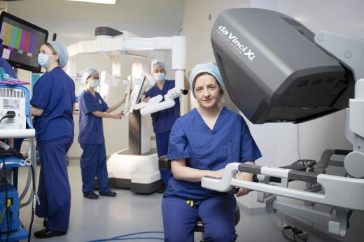 A doctor sitting down next to a da Vinci Xi robot, with a team of doctors behind her in blue overalls and masks working robotic equipment