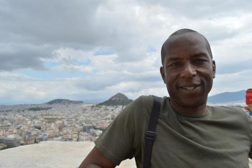 A man smiling, with an elevated view of a city behind him and mountains in the background