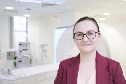 A doctor dressed smartly in a dark pink suit, wearing glasses and hair tied back, standing in front of a white machine like looks like an MRI